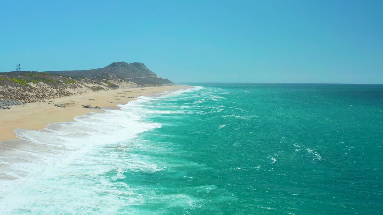 At 0:00, La Laguna Beach in Los Cabos Mexico appears from above with turquoise surf curling onto pale sand, rocky dunes in the distance, blue sky, and no visible people.
