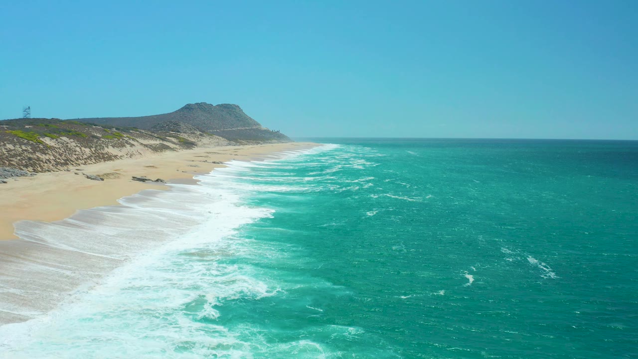 At 0:02, La Laguna Beach fills the left side of the frame while bands of white surf break beside turquoise water, a rocky headland, clear sky, and no visible people.