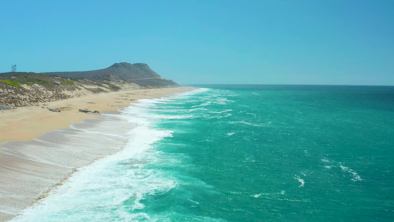 Aerial view of foamy surf rolling onto empty sand with turquoise ocean beside it.