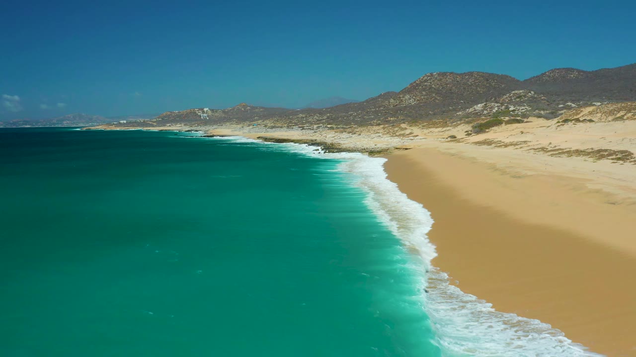 White waves curl along a pale beach below rocky mountains