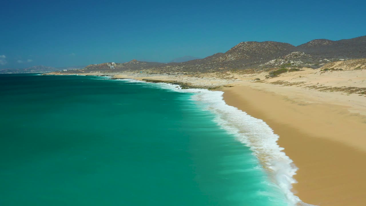 Aerial view over La Laguna Beach, with a curved foam edge meeting smooth sand, broad turquoise water in the foreground, desert dunes behind the beach, and no visible people.