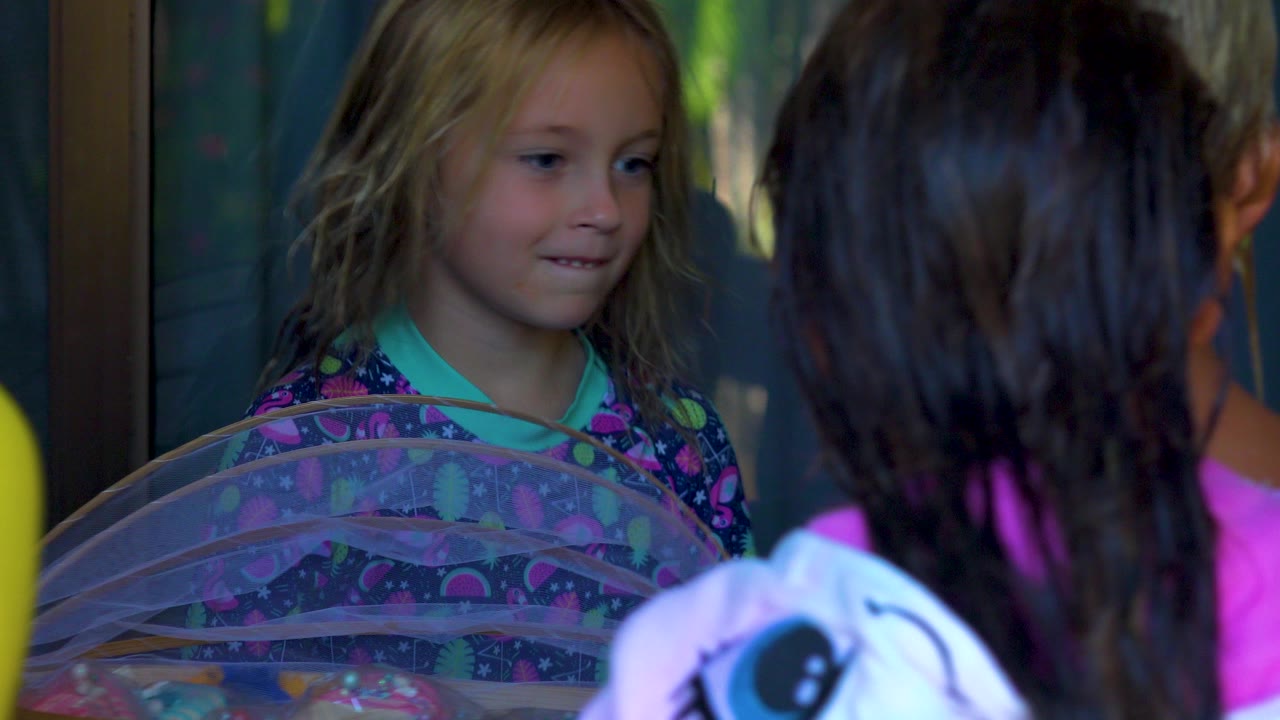 A blonde girl in a colorful shirt faces other children near a translucent covered table in San Jose del Cabo, Mexico, with glass doors and greenery behind her.