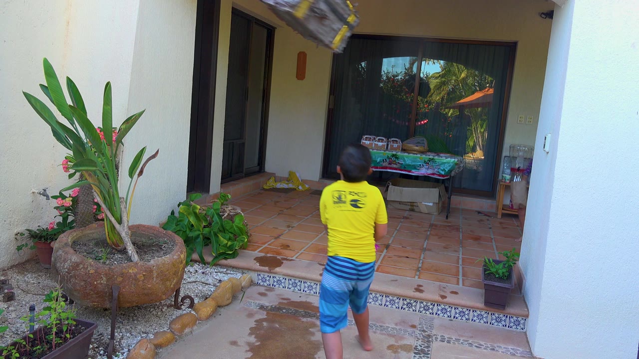 A child in a yellow shirt and blue shorts stands on a tiled patio in San Jose del Cabo BCS Mexico, facing a hanging pinata near potted plants and glass doors.