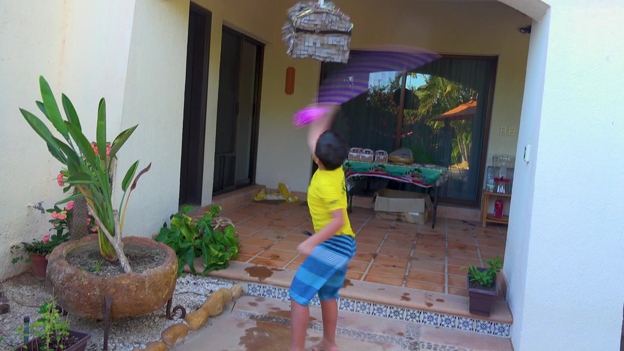 A child in yellow and blue swings a purple stick upward at a hanging pinata on a tiled patio in San Jose del Cabo BCS Mexico, with plants and glass doors behind.