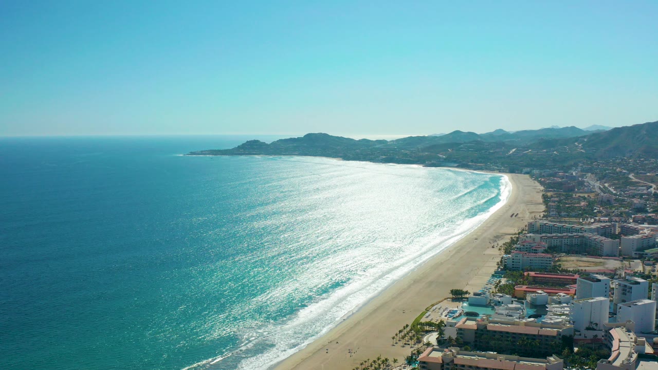 Aerial view of turquoise ocean, sandy beach, coastal buildings, and distant hills.