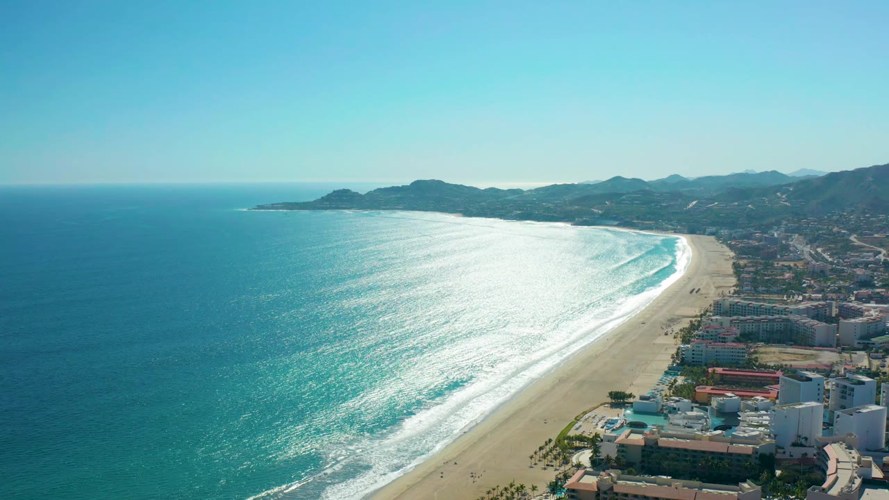 Aerial view in San Jose del Cabo, Mexico with the ocean dominating the left frame, white waves reaching the sand, beachfront buildings at lower right, distant hills, and no visible people.