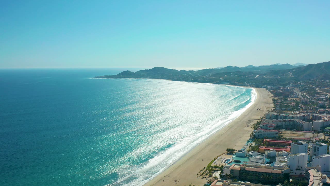San Jose del Cabo, Mexico aerial frame showing a broad turquoise bay, a curving sandy beach, white surf bands, shoreline buildings, low mountains, and no visible people.