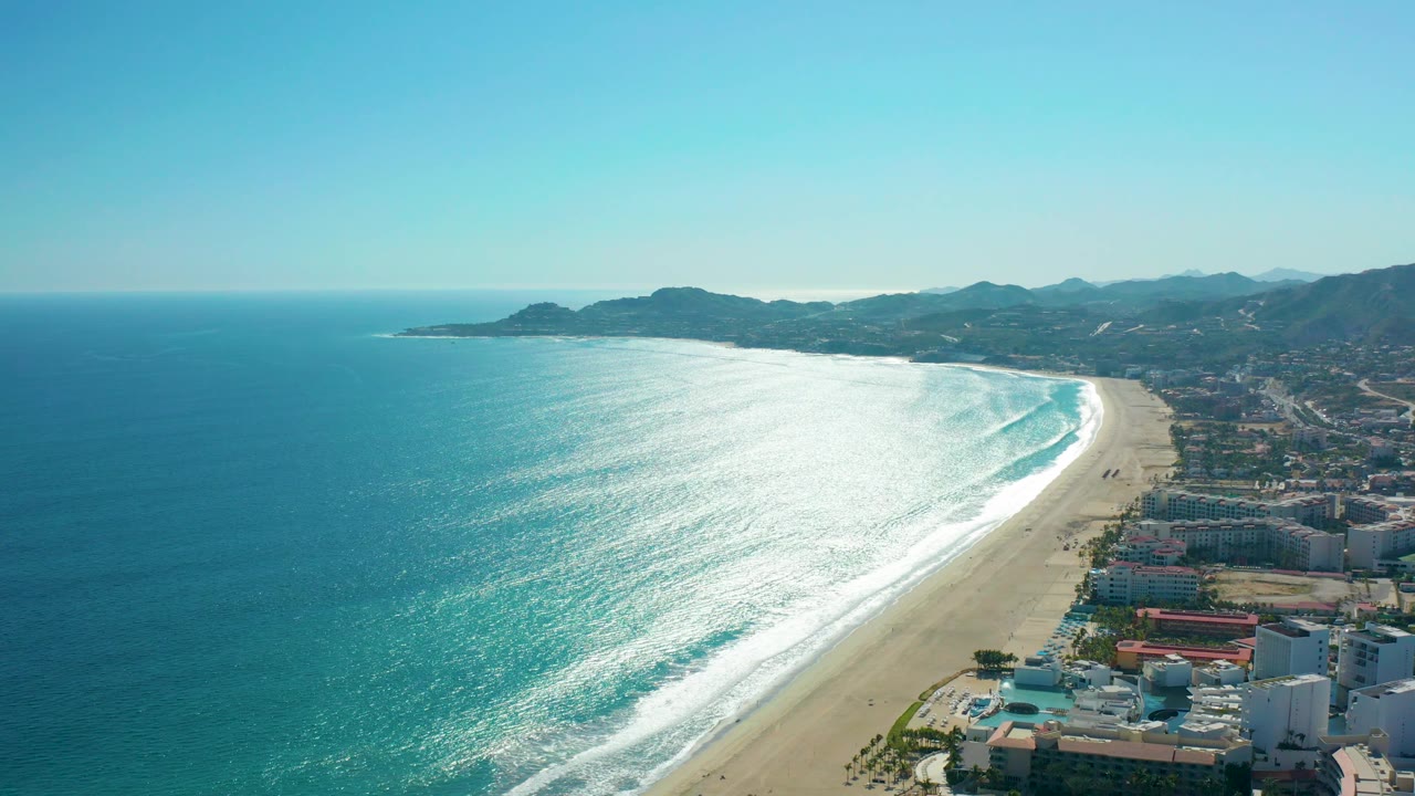 Aerial frame over San Jose del Cabo, Mexico with more open water at left, a bright surf edge along the beach, clustered coastal buildings to the right, hills in back, and no visible people.