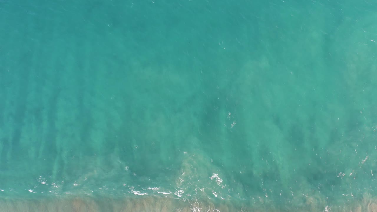 Aerial view of turquoise water in San Jose del Cabo BCS Mexico, where soft sand streaks show through the surface and small white wave flecks appear near the lower edge, with no visible people.