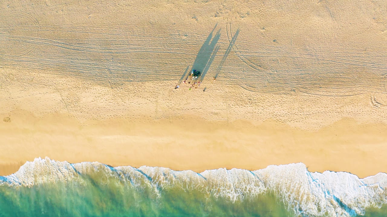 Overhead view of Diamante Cabo San Lucas beach in Mexico, with white surf at the lower edge and small visible people casting long shadows across pale sand near the top.