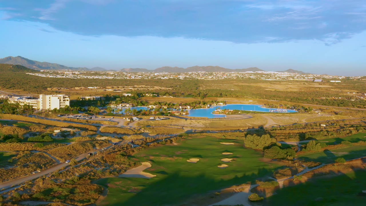 Aerial view in Mexico with golf greens and bunkers in the foreground, a bright pool and building near the center, distant development on hills, and no visible people.
