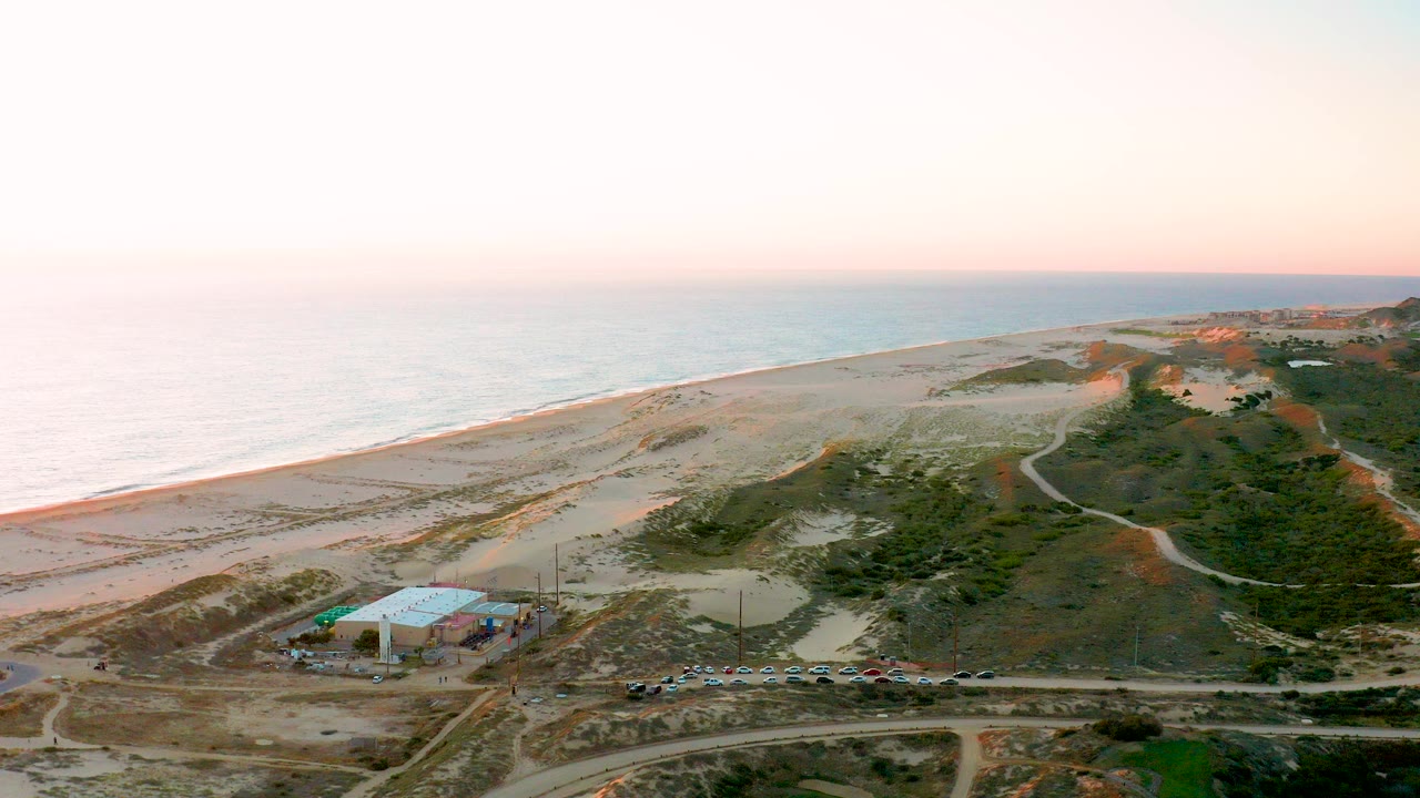 Aerial view of sandy beach, ocean, dunes, road, parked cars, and a blue-roofed building.