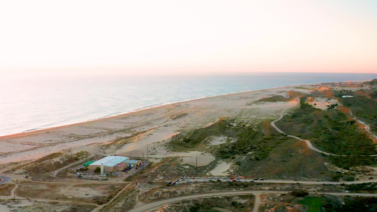 A wide aerial view in Diamante Cabo San Lucas BCS Mexico frames the pale beach and ocean behind dunes, dirt tracks, parked vehicles, and the blue-roofed structure, with no visible people.