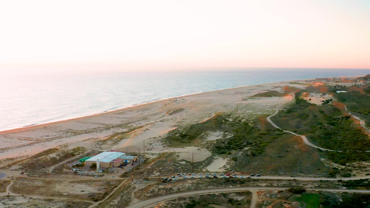 Drone-style aerial frame in Diamante Cabo San Lucas BCS Mexico showing coastal dunes between the ocean and a dirt road lined with parked cars near a blue-roofed building, with no visible people.