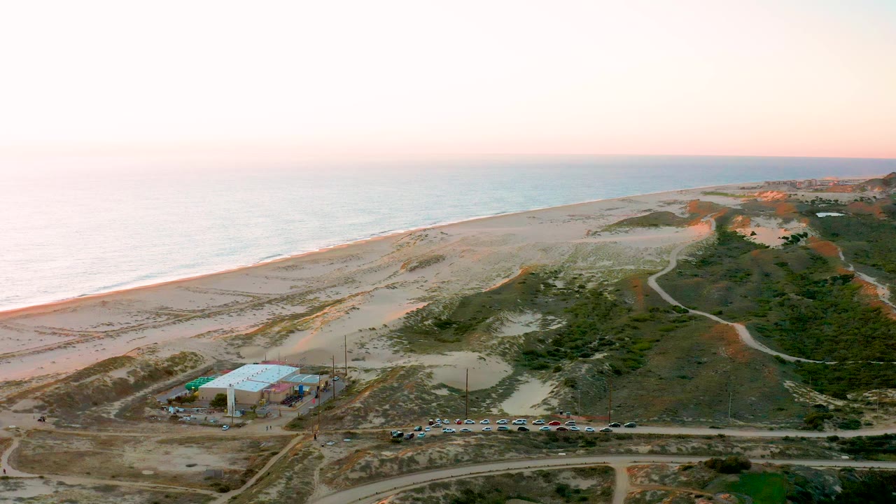 Aerial frame over Diamante Cabo San Lucas BCS Mexico with hazy ocean light, a long sandy beach, scrub-covered dunes, a road, parked cars, and the blue-roofed building, with no visible people.