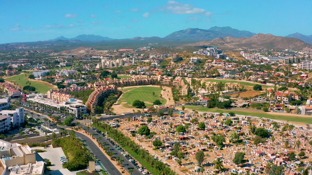 Opening aerial frame over San Jose del Cabo with a foreground road and cemetery-like lots, a green golf course in the midground, residential buildings beyond, distant mountains, and no visible people.
