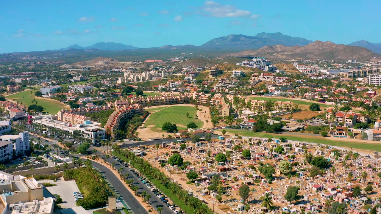 Early aerial view in San Jose del Cabo showing cars along a curving road, a broad green golf course, clustered housing, pale buildings, dry foothills, distant mountains, and no visible people.