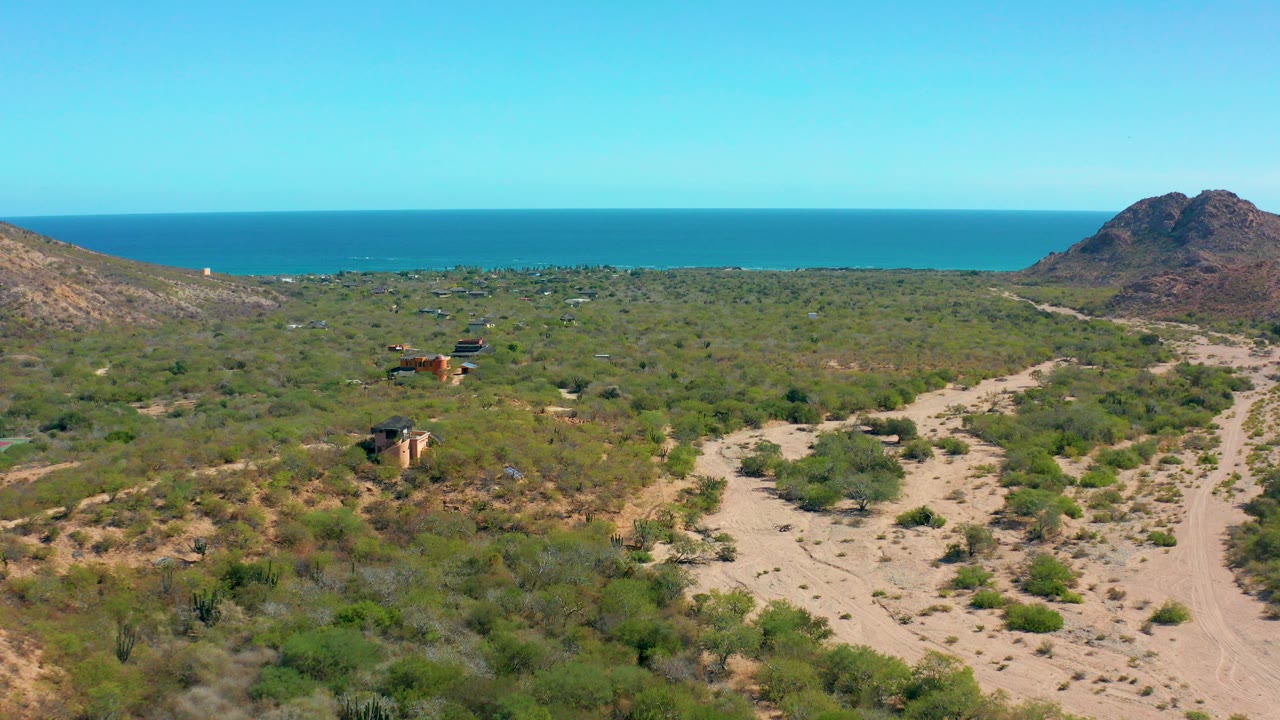 Aerial view in Cabo Pulmo, Mexico with sandy tracks crossing green scrub toward a reddish hill and blue ocean horizon, with no visible people.