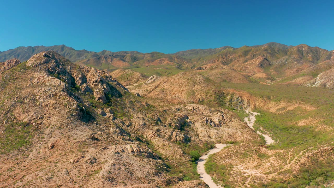 Aerial view in Cabo Pulmo BCS Mexico with a curving dirt track in the lower valley, rugged tan slopes, scattered green vegetation, mountains across the horizon, and no visible people.