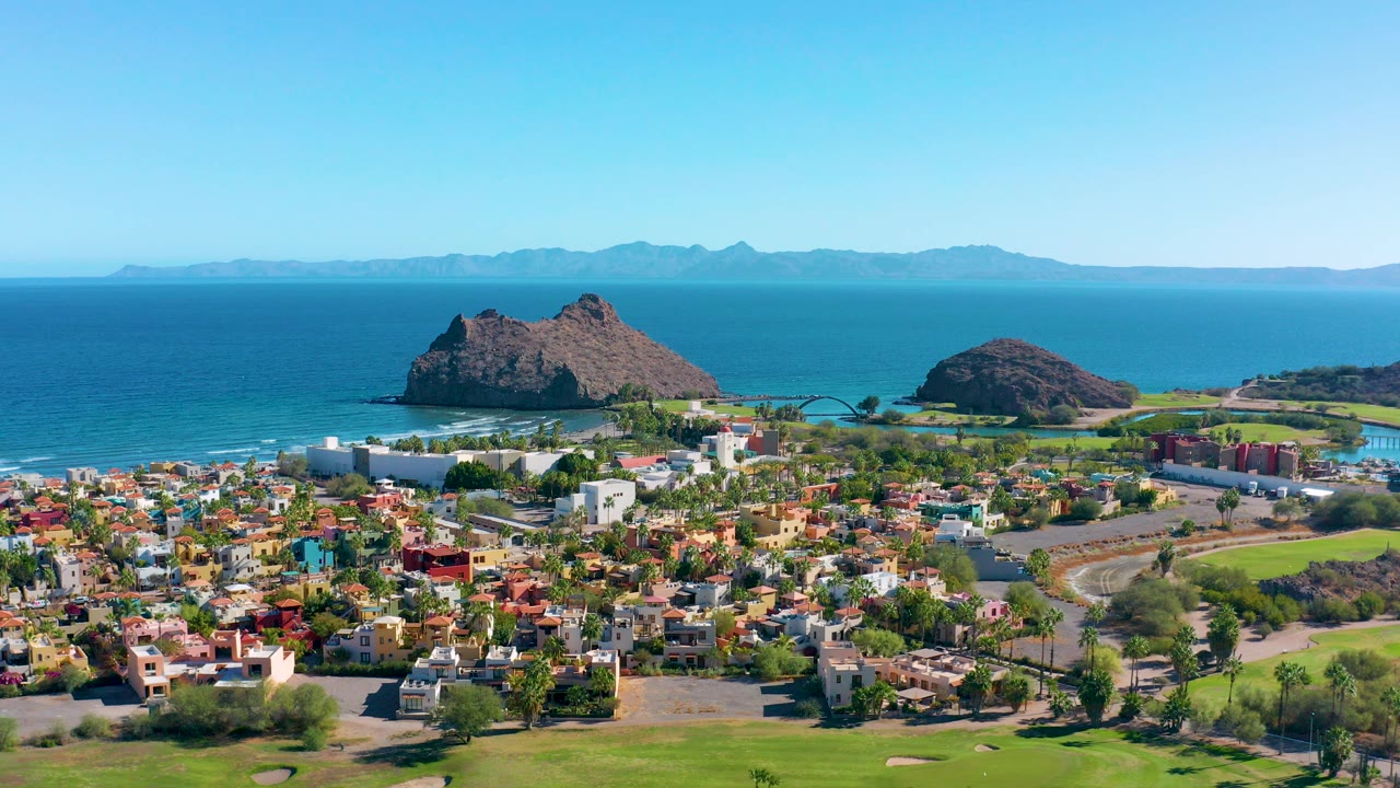 Opening aerial frame over Loreto BCS Mexico with colorful town blocks in the foreground, rocky outcrops near the shoreline, blue ocean, distant mountains, and no visible people.