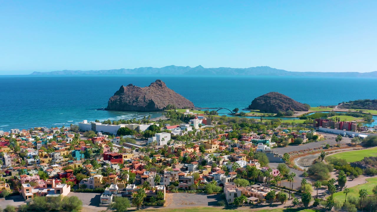 Aerial coastal town with low buildings, white structures, green fields, rocks, and sea