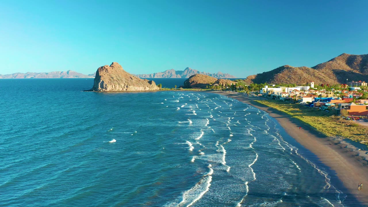 At 0:01, the Loreto BCS Mexico shoreline remains in a wide aerial composition, with whitecaps slightly farther along the beach, buildings at right, rocky hills beyond, and no clearly visible people.