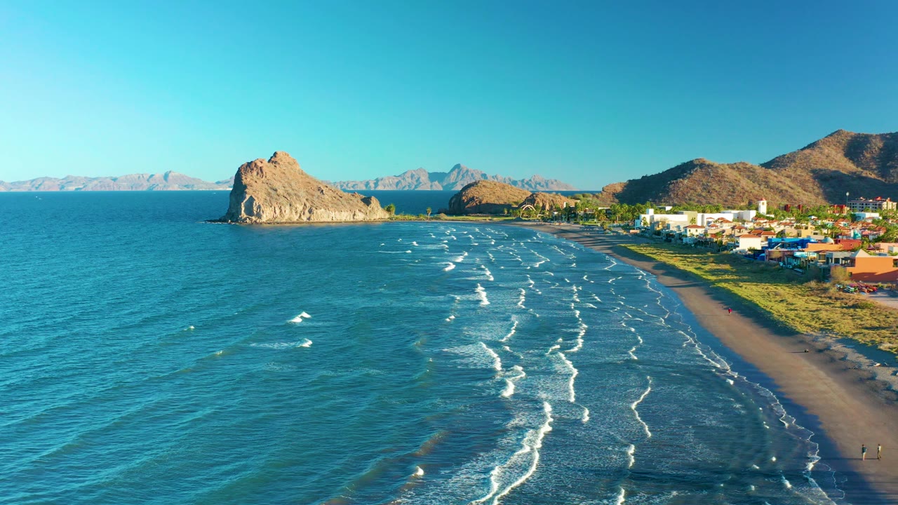 At 0:03, the aerial view over Loreto BCS Mexico keeps the beach on the right edge, with small white waves across the water, low coastal buildings, sunlit hills, distant mountains, and no clearly visible people.