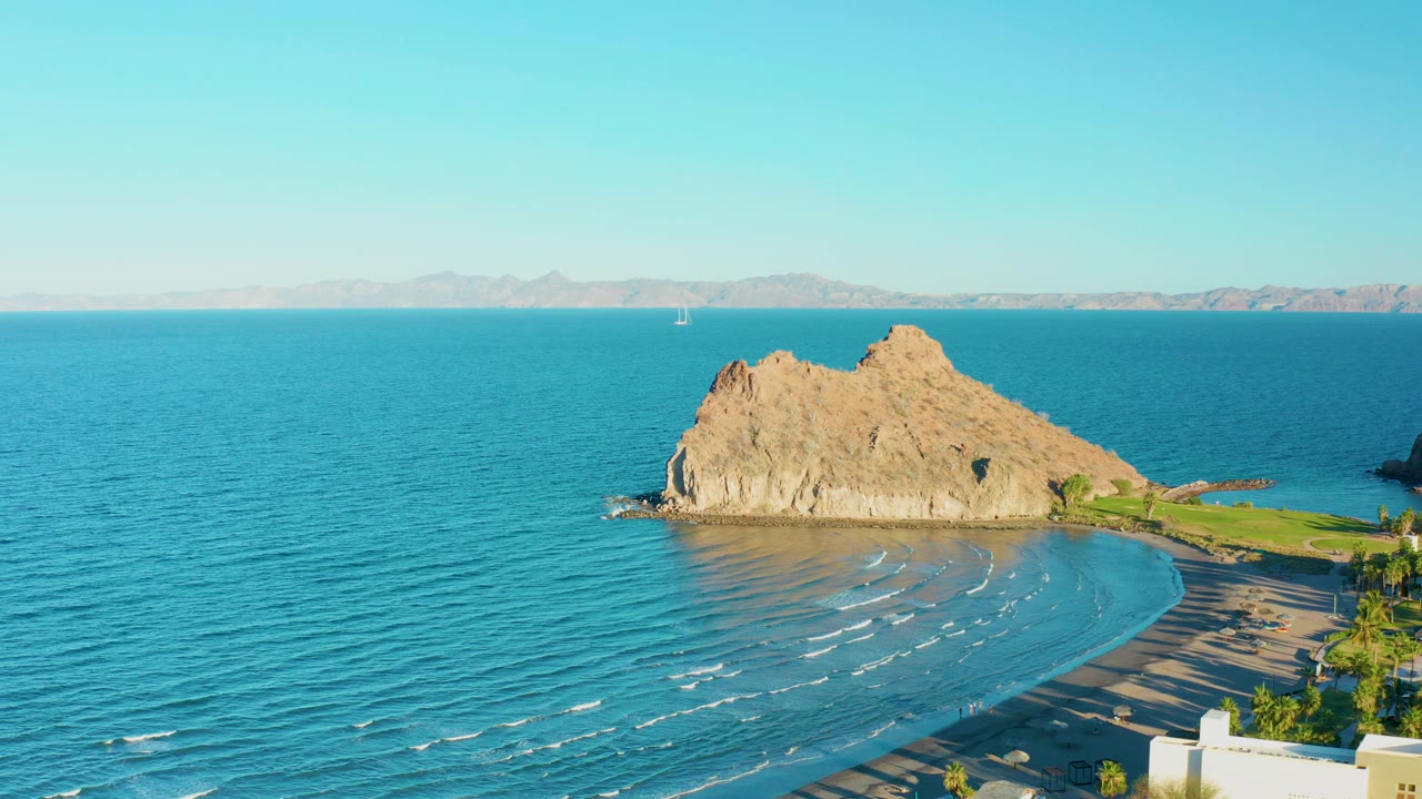 Loreto BCS Mexico aerial frame with a tan rocky outcrop beside turquoise water, a curved sandy beach, white foreground buildings, distant mountains, and no visible people.