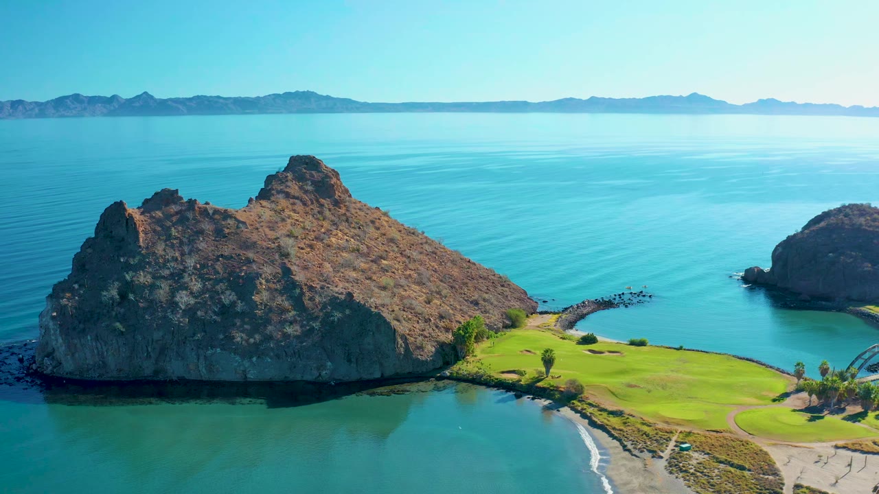 Aerial view in Loreto BCS Mexico with the rocky island centered left, calm blue water behind it, a green coastal edge below, and no visible people.