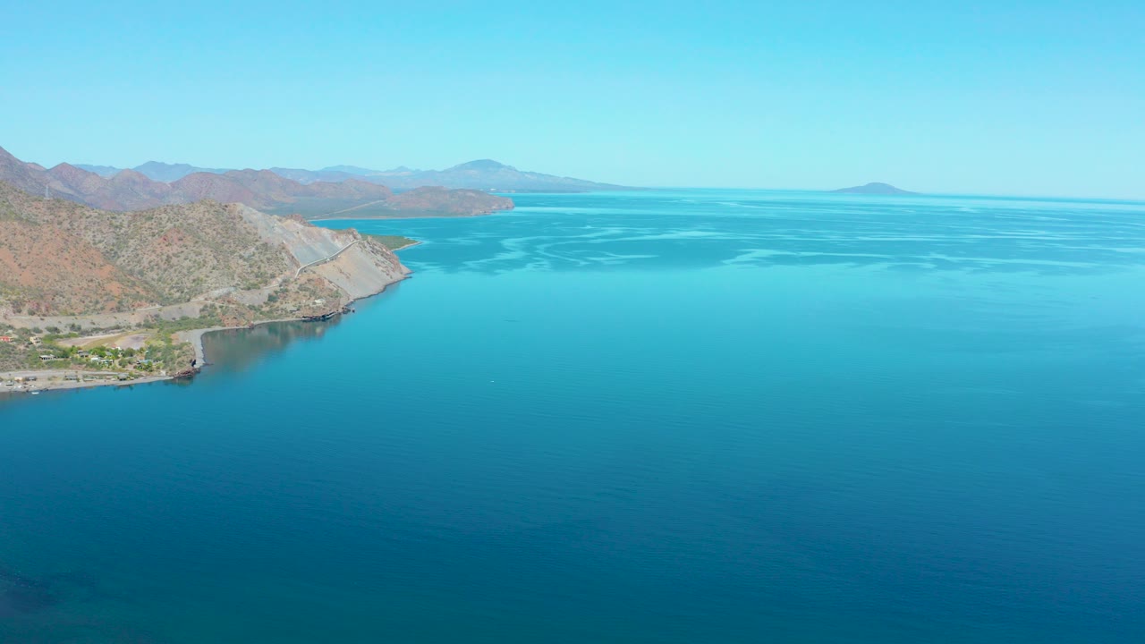 Aerial view in Loreto BCS Mexico showing a rocky shoreline at left, turquoise water across the frame, distant mountains, pale sky, and no visible people.