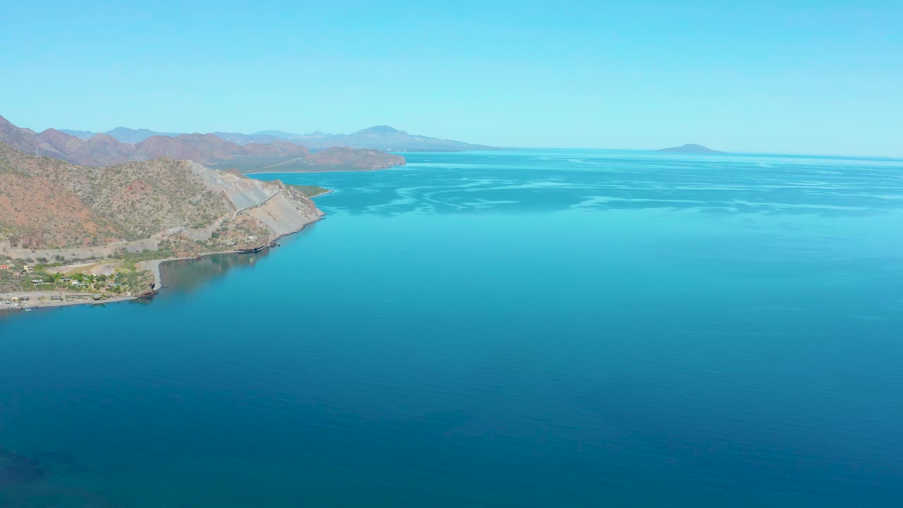 Wide aerial frame of Loreto BCS Mexico with the shoreline occupying the left edge, bright turquoise shallows, darker open water, distant mountains, and no visible people.