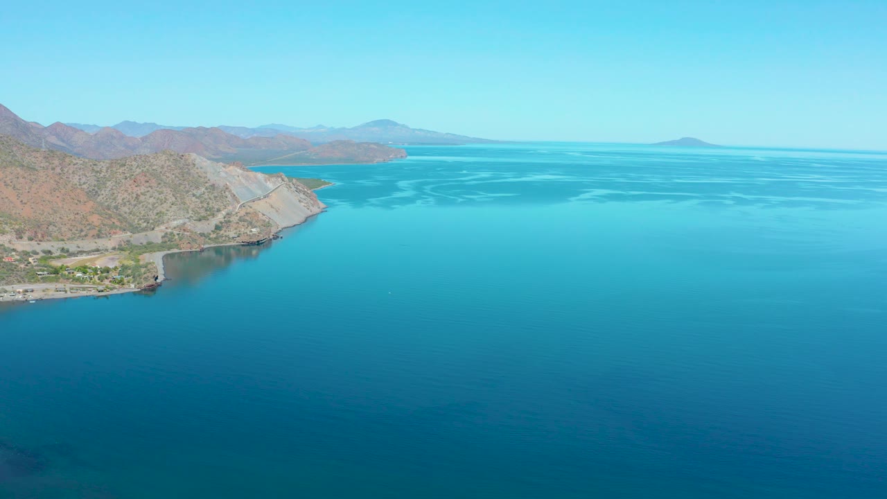 Aerial frame in Loreto BCS Mexico with a rocky point and tan mountain slope on the left, broad turquoise coastal water to the right, distant landforms, and no visible people.
