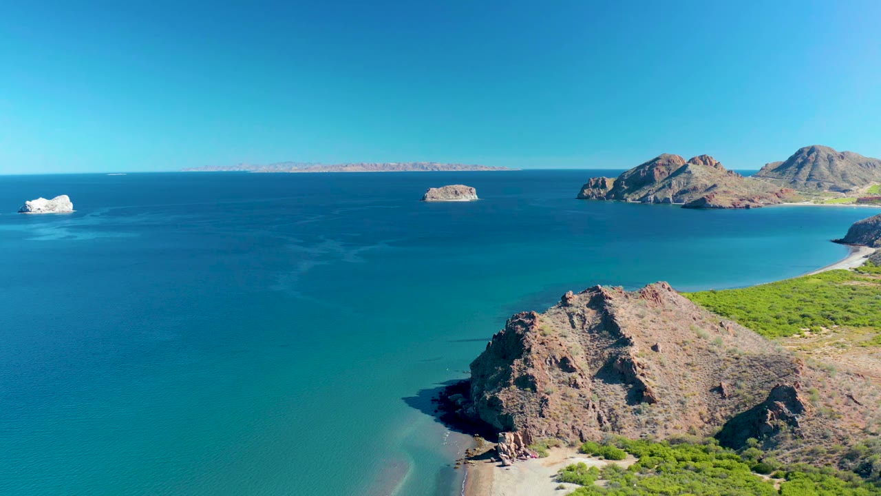 High aerial frame of Ligui BCS Mexico showing the foreground cliff slightly lower in frame, the broad turquoise bay, small offshore rock forms, and no visible people.