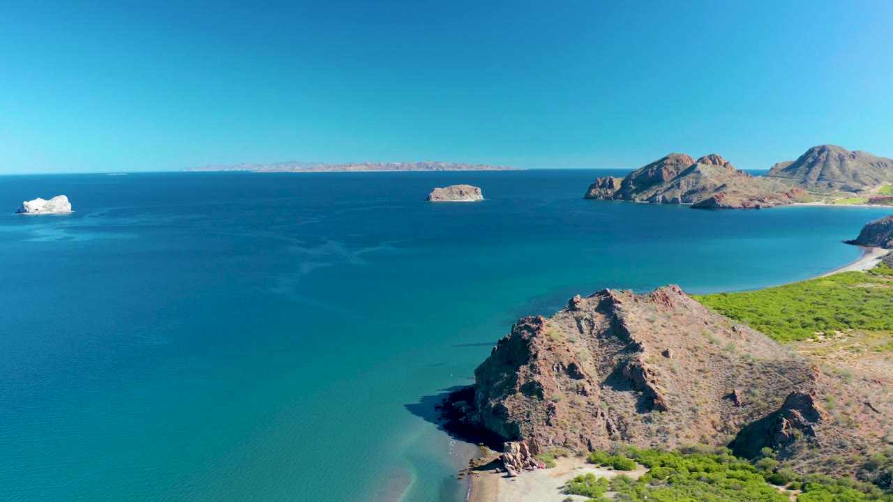 Ligui BCS Mexico coastline seen from above, with a rocky point at lower right, shallow turquoise water near the beach, distant island rocks, and no visible people.