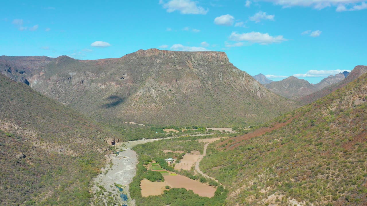 Wide aerial view in San Javier BCS Mexico showing a pale riverbed at lower left, green valley vegetation, brown hills, flat-topped mountains, scattered clouds, and no visible people.