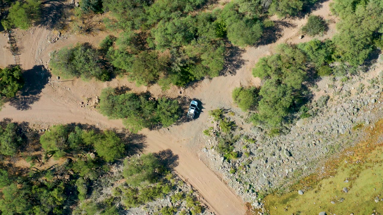Overhead frame from San Javier BCS Mexico showing a white car on a pale dirt road with green trees, rocky ground, hard shadows, and no visible people.