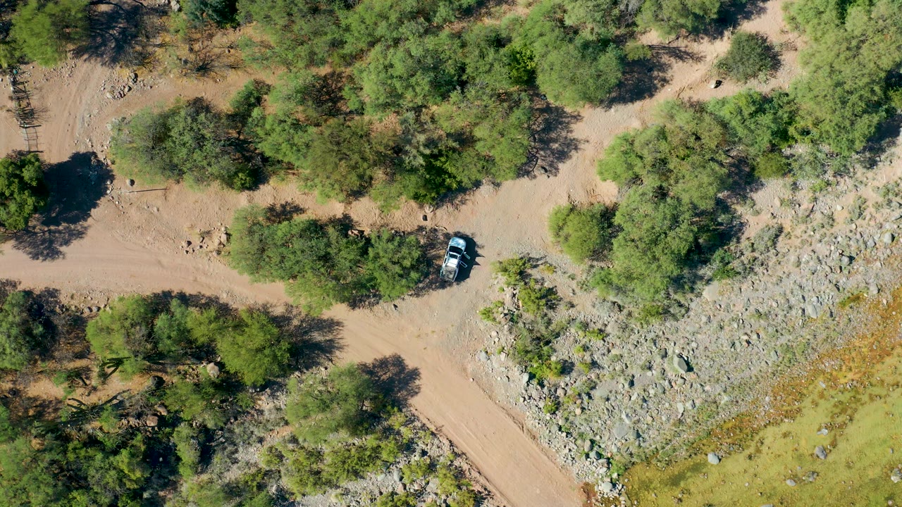 High aerial frame in San Javier BCS Mexico showing the white car near a cluster of trees on a dusty road, with a rocky wash to one side and no visible people.