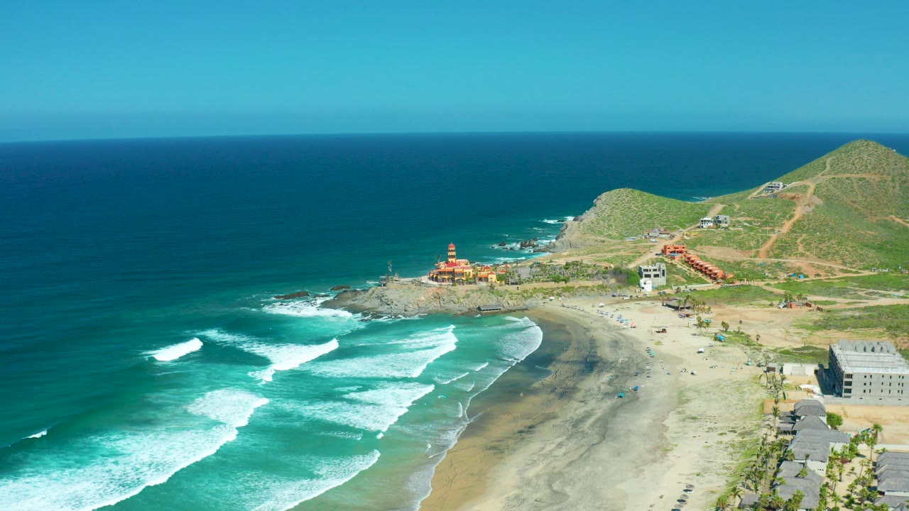 Aerial frame over Cerritos Beach BCS Mexico with white surf rolling toward a sandy curve, blue ocean at left, hillside buildings beyond the rocky point, and no visible people.