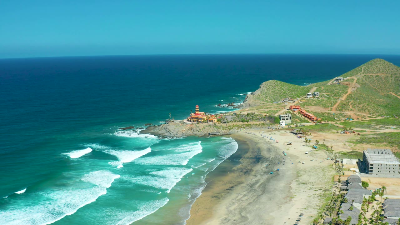 A wide aerial view of Cerritos Beach BCS Mexico with bright surf lines on the left, a pale sandy beach below, hillside paths and buildings to the right, and no visible people.