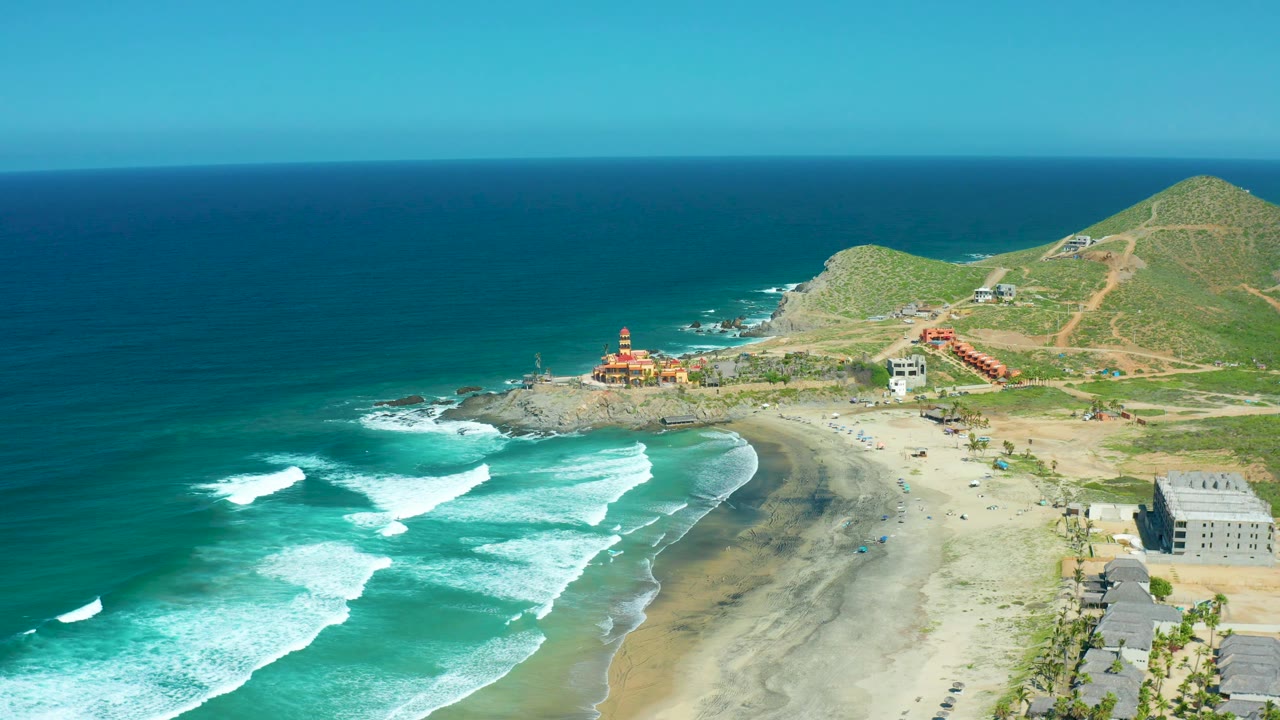 Ocean waves break diagonally toward a sandy Mexican beach with hills behind.