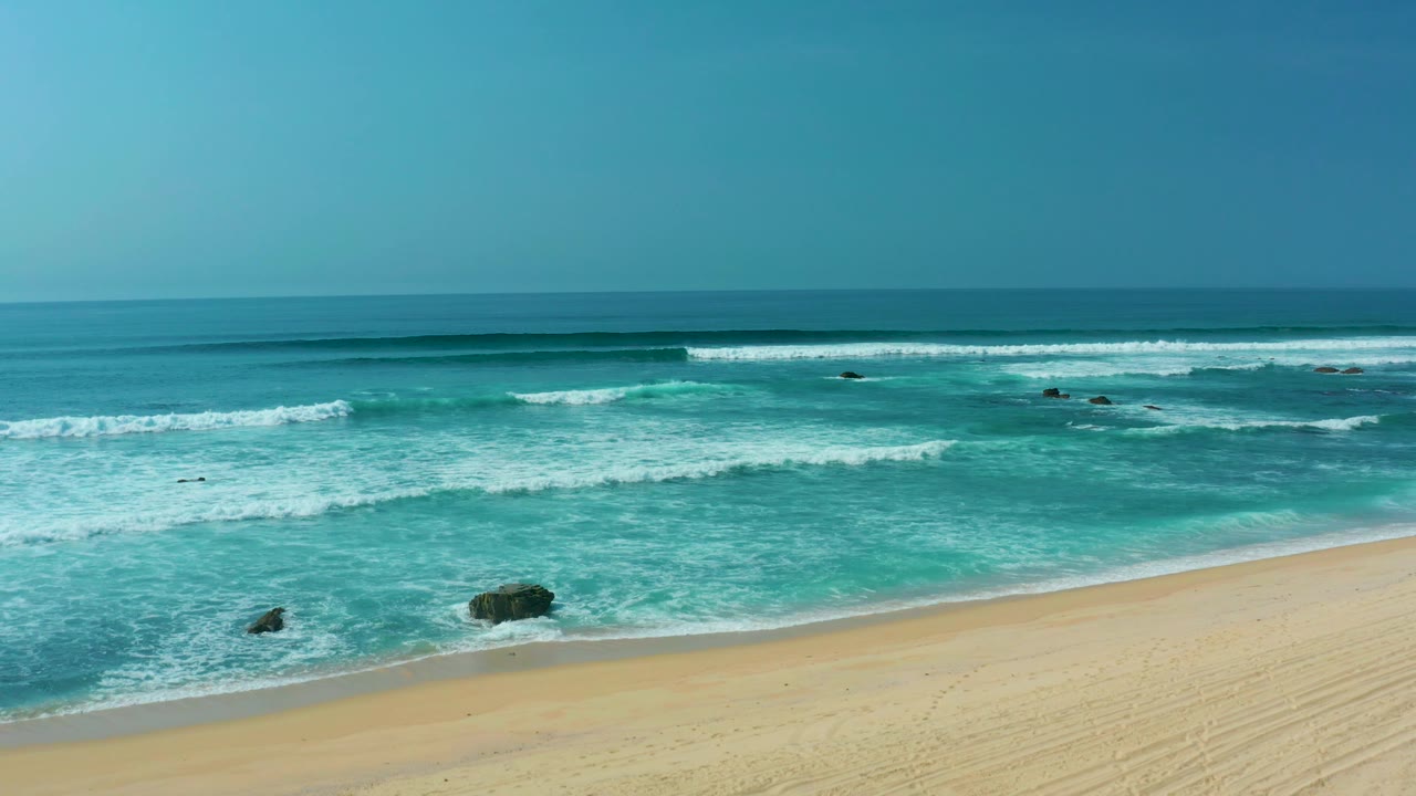 Opening frame of San Luis Beach, East Cape, Los Cabos, Mexico, with turquoise surf breaking toward pale sand, dark rocks in shallow water, and no visible people.