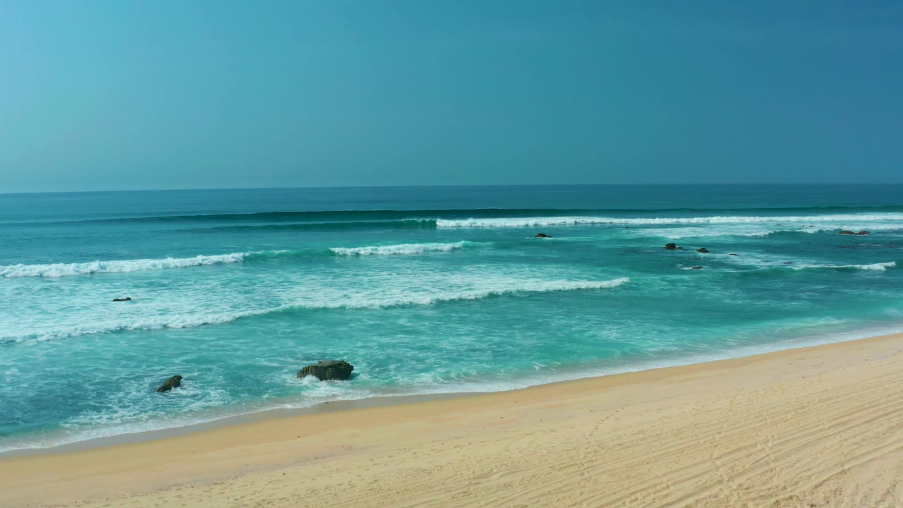 A later moment at San Luis Beach shows waves rolling over scattered dark rocks toward empty sand, with the water shifting from deep blue to turquoise and no visible people.