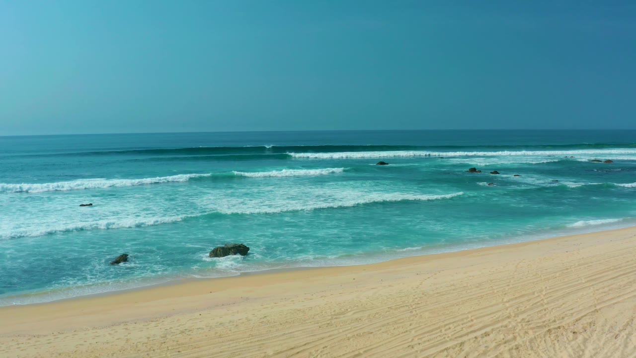 San Luis Beach remains empty as white foam approaches the sand, with several dark rocks visible in shallow turquoise water and no visible people.
