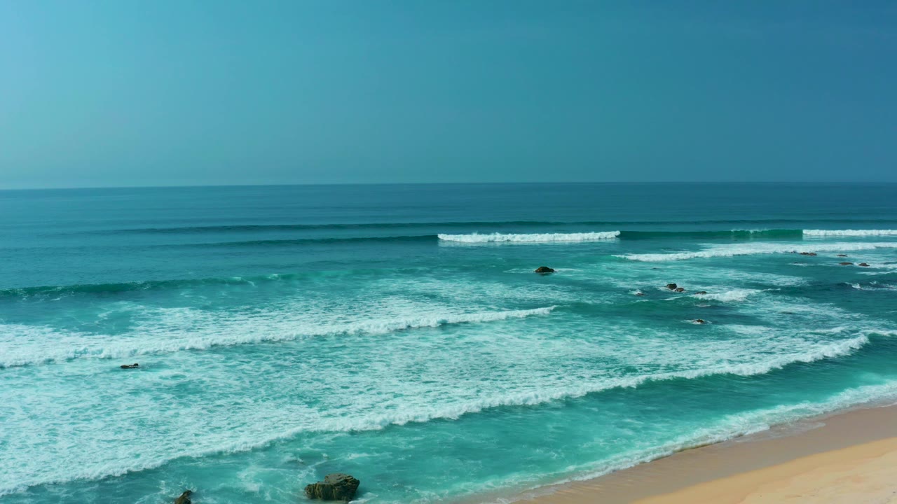 Turquoise waves breaking toward a sandy beach under a clear sky.