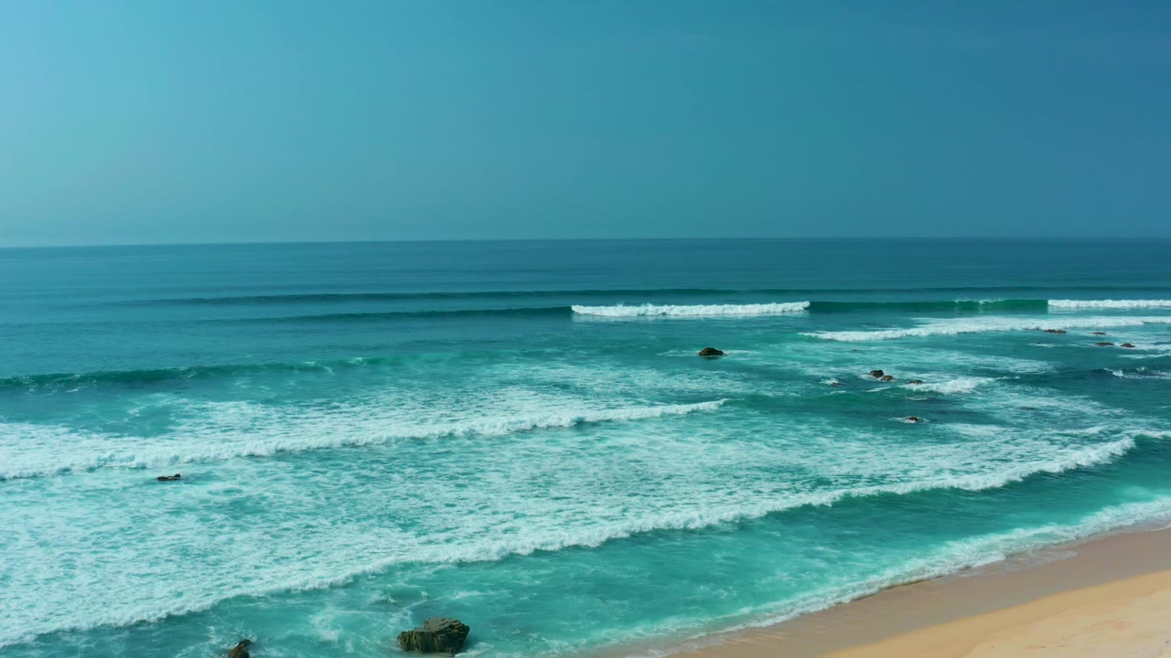 San Luis Beach aerial frame showing white foam spreading across shallow turquoise water, dark rocks offshore, sandy beach at the lower right, and no visible people.