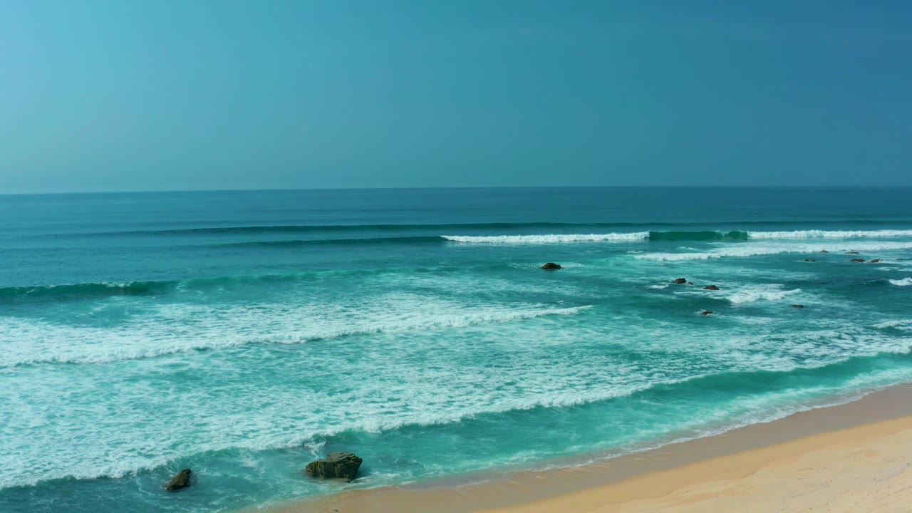 San Luis Beach coastline seen from above with a level blue horizon, breaking surf across the center, dark rocks near shore, sandy beach below, and no visible people.