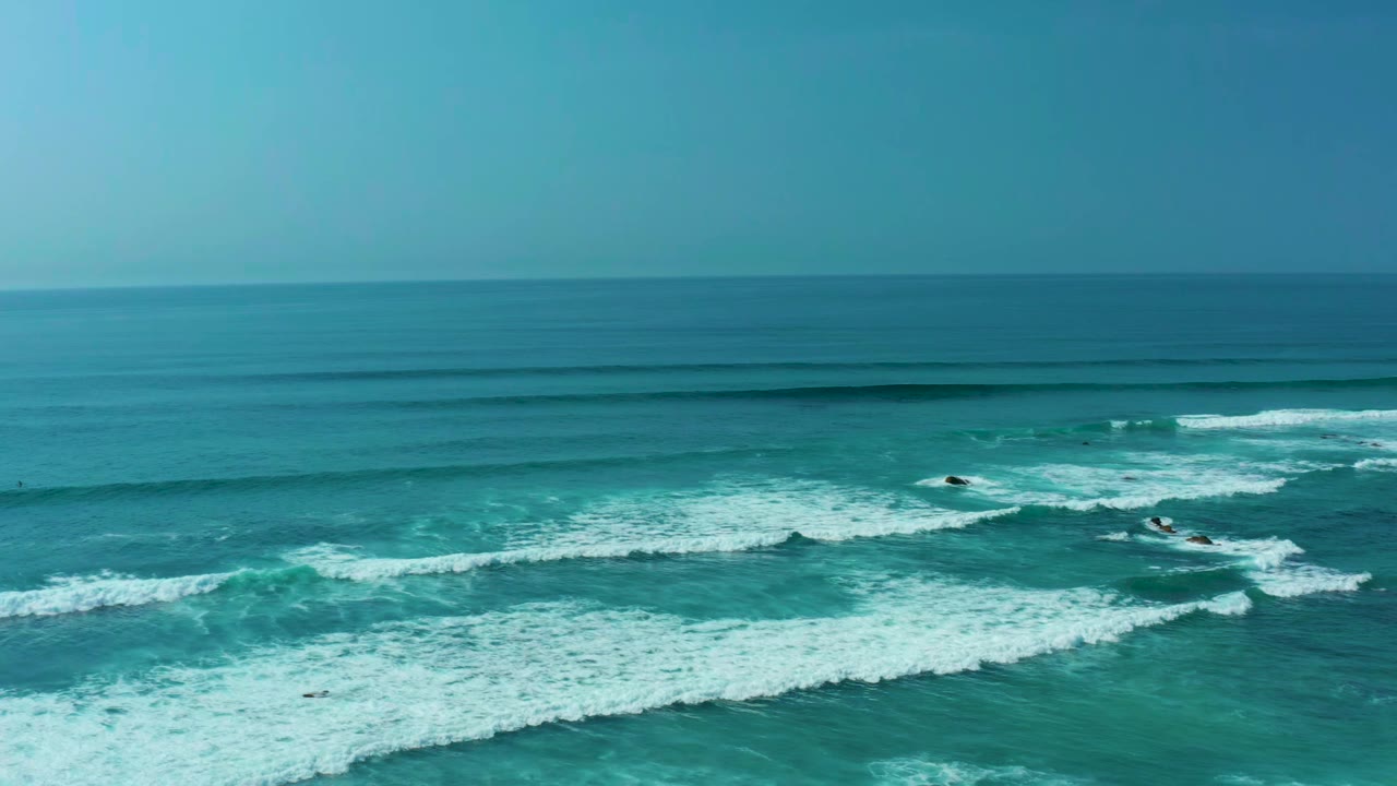 Aerial frame from San Luis Beach, East Cape Los Cabos showing turquoise water, white surf bands, dark offshore rocks, a straight horizon, and no visible people.