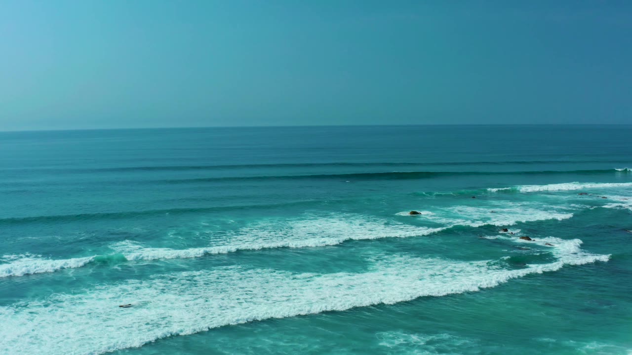San Luis Beach, East Cape Los Cabos appears as a wide ocean composition with white foam curling around dark offshore rocks, open blue water, a level horizon, and no visible people.