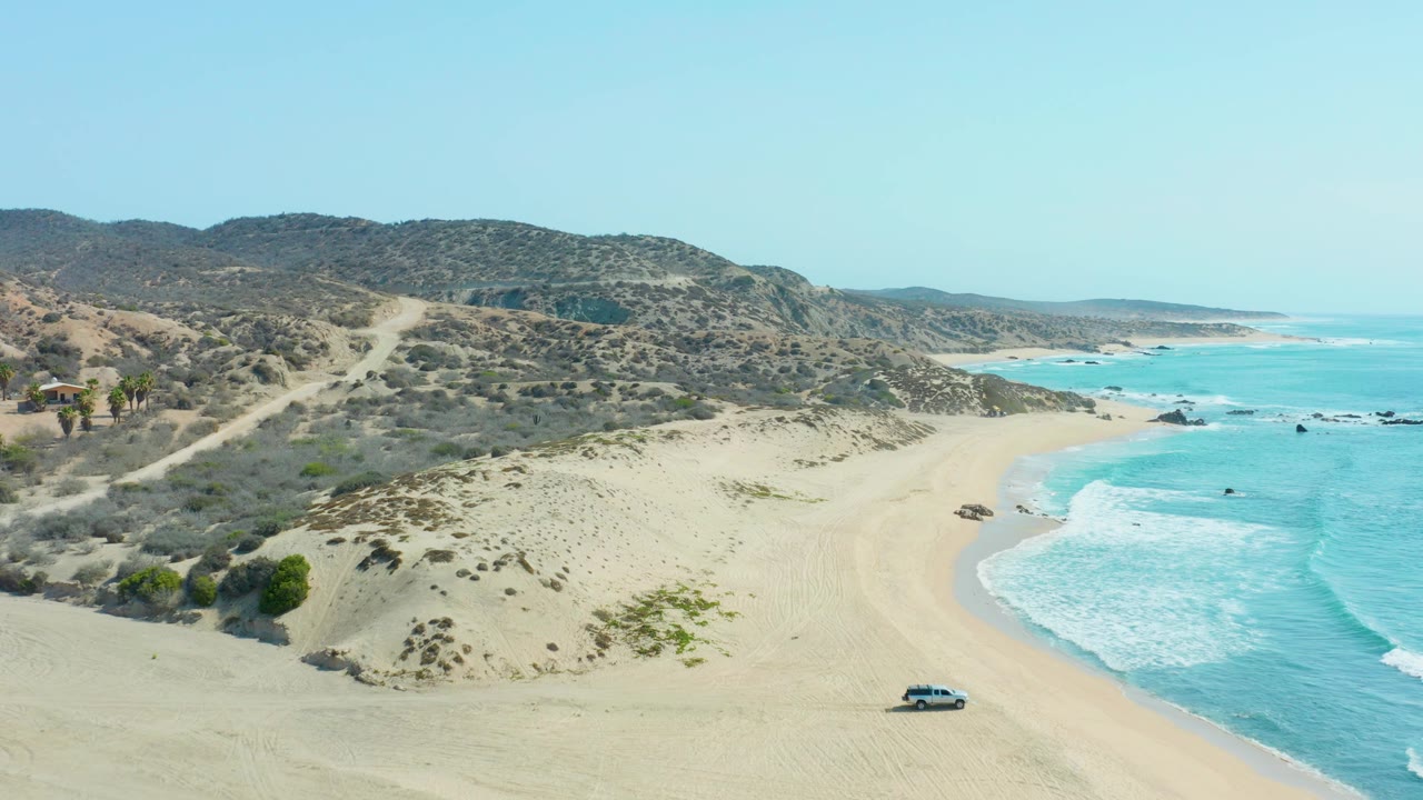 Aerial view over San Luis Beach East Cape Los Cabos with the vehicle near the lower sand, foamy waves to the right, dune ridges in midground, and no visible people.