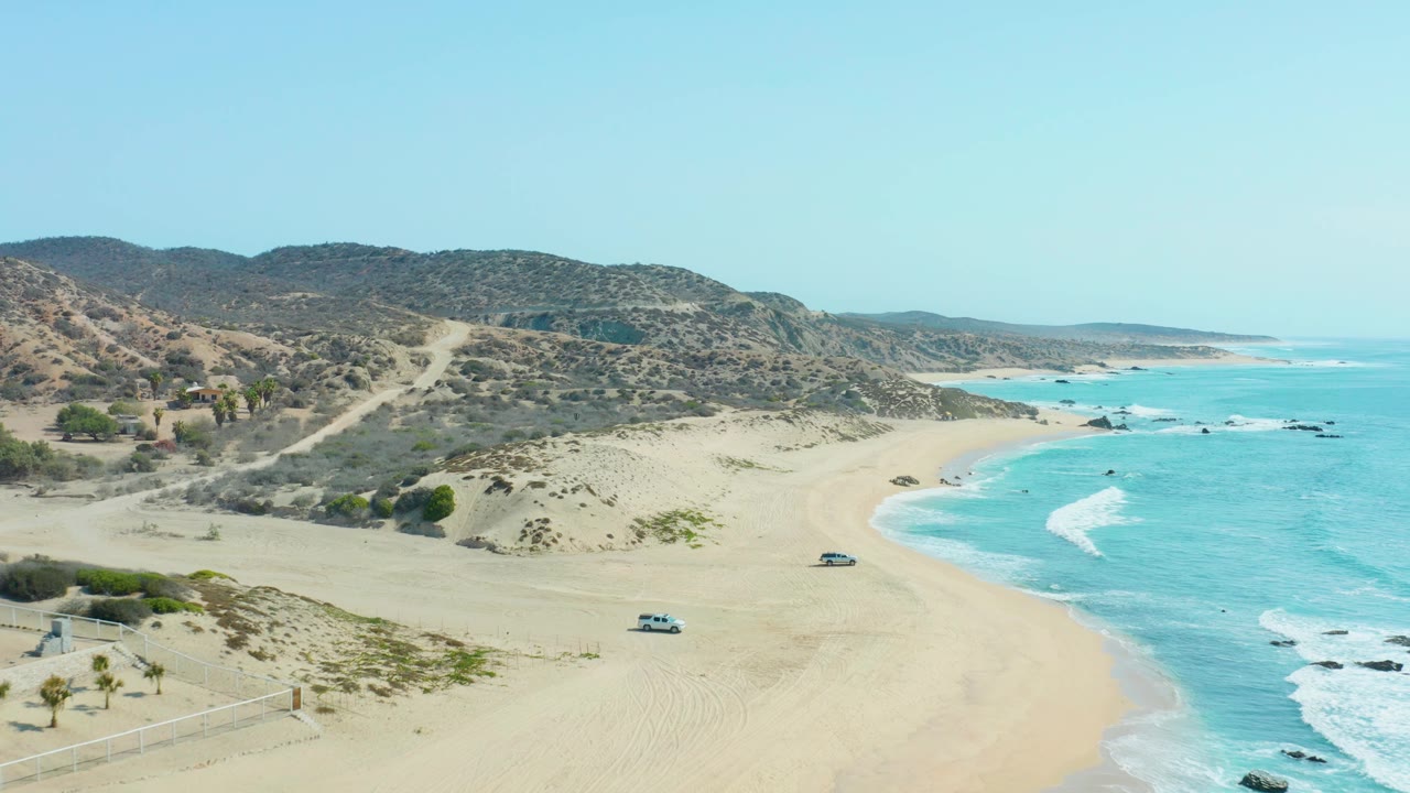 San Luis Beach East Cape Los Cabos appears from above with vehicles spaced across the sand, waves curling along the right side, and no visible people.